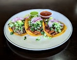 Three tacos placed on a white plate, filled with a variety of ingredients including chopped lettuce, diced onions, and slices of red onion. Each taco contains different types of meat, and they are accompanied by two small cups of sauce, one green and one red. A lime wedge is also present on the plate.