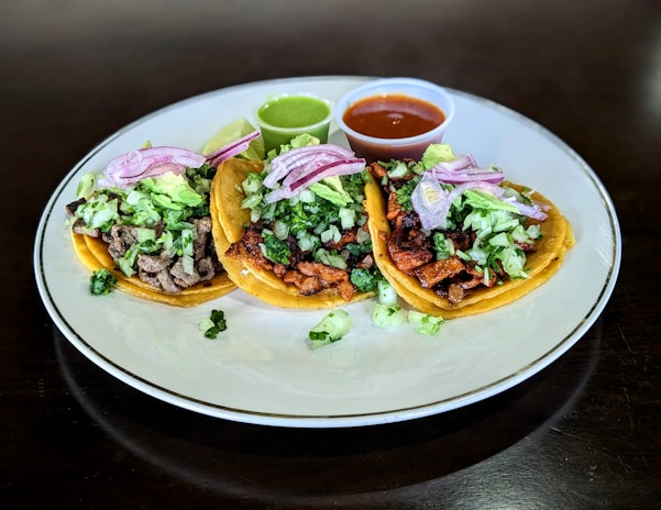 Three tacos placed on a white plate, filled with a variety of ingredients including chopped lettuce, diced onions, and slices of red onion. Each taco contains different types of meat, and they are accompanied by two small cups of sauce, one green and one red. A lime wedge is also present on the plate.