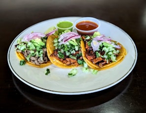 Three tacos placed on a white plate, filled with a variety of ingredients including chopped lettuce, diced onions, and slices of red onion. Each taco contains different types of meat, and they are accompanied by two small cups of sauce, one green and one red. A lime wedge is also present on the plate.