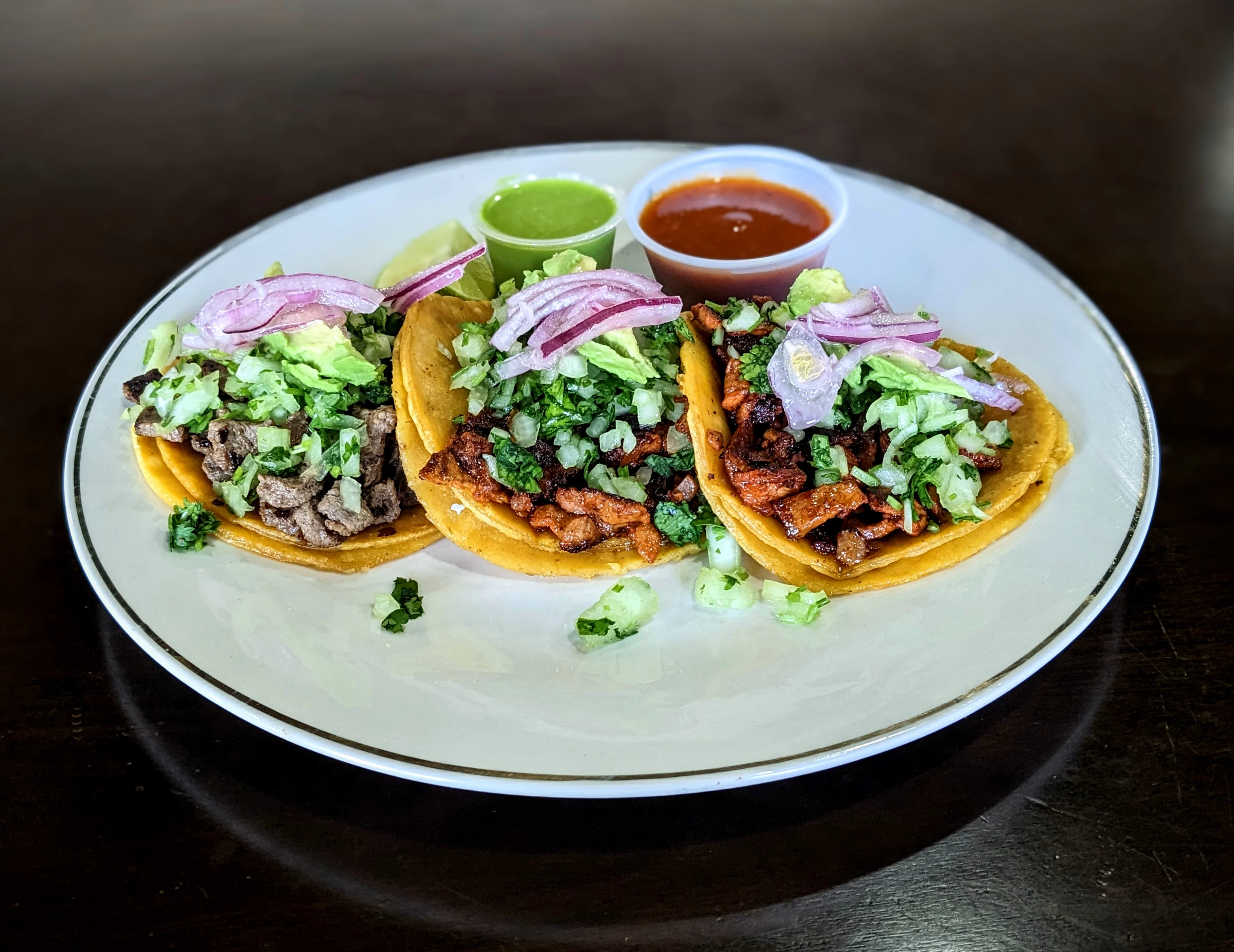 Three tacos al pastor topped with onion, cilantro and pickled red onion, with green and red salsas