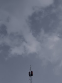A tall telecommunications tower extends upwards into a cloudy sky, with a few clouds scattered around, and features a metal structure with antennas.