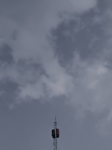 A tall telecommunications tower extends upwards into a cloudy sky, with a few clouds scattered around, and features a metal structure with antennas.