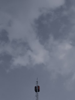 A tall telecommunications tower extends upwards into a cloudy sky, with a few clouds scattered around, and features a metal structure with antennas.