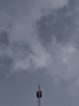 A tall telecommunications tower extends upwards into a cloudy sky, with a few clouds scattered around, and features a metal structure with antennas.