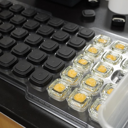 A collection of clear plastic containers holding small amounts of orange-colored substance is arranged on a tray. Adjacent to it, there is a grid of black, square spaces or buttons. The setting appears to be a laboratory or testing environment with a focus on organization and controlled conditions.