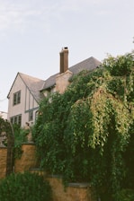 A two-story house with a steeply pitched roof and a prominent chimney. The house exterior features a combination of stone and light-colored stucco with decorative trim. Lush green foliage and trees partially obscure the lower part of the house, and a stone wall with a metal archway is visible in the foreground.