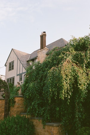 A two-story house with a steeply pitched roof and a prominent chimney. The house exterior features a combination of stone and light-colored stucco with decorative trim. Lush green foliage and trees partially obscure the lower part of the house, and a stone wall with a metal archway is visible in the foreground.