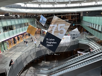 An interior view of an airport featuring escalators, people walking, and large hanging signs celebrating a 75th anniversary. The area is spacious with a modern design, including glass railings and open spaces. The scene is bustling with travelers and decorated with banners.