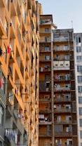 A cluster of high-rise apartment buildings with balconies. There are various clothes hanging out to dry, indicating a residential area. The buildings have a mix of grey and orange hues, with signs of daily life visible through open windows and balconies.