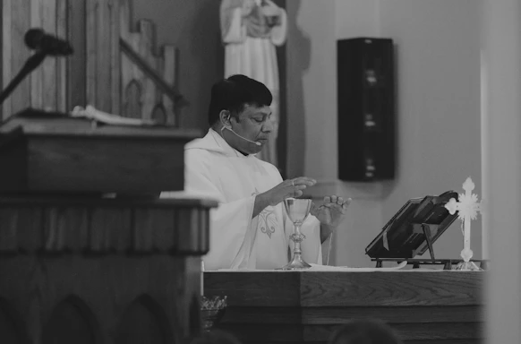 A person dressed in religious vestments appears to be conducting a ceremony. A chalice is placed on an altar, and the individual looks focused and engaged. A microphone is visible, suggesting the ceremony might be being amplified for an audience. Religious symbols and a wooden structure are visible in the background.
