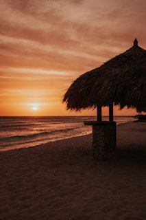 A serene beach scene at sunset with an orange and pink hued sky. The silhouette of a thatched-roof hut on stilts stands prominently on the sand near the water's edge. The ocean waves gently lap the shore, creating a calm and peaceful ambiance.