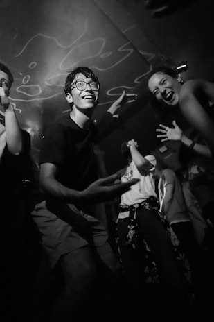 A group of young people engaging in lively and enthusiastic dancing within a dimly lit environment, featuring dynamic expressions of joy and movement. The scene captures a sense of fun and camaraderie.
