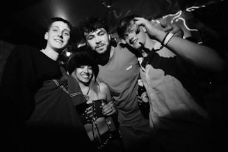 A group of four young adults are gathered in a dimly lit room, possibly at a party or social gathering. They are all smiling and appear to be enjoying themselves. Two of them are holding drinks. The image is in black and white, which gives it a classic and timeless feel.