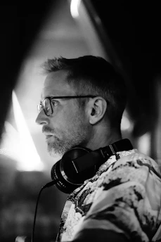 A black and white image of a person wearing glasses and headphones, focused intently to the side. The environment appears dimly lit, casting shadows and highlights that create a moody atmosphere.