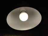 Close-up of a ceiling light fixture with a round white bulb emitting bright light. The fixture has a white inner surface creating a contrast against the dark background.