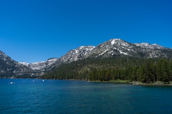 A serene landscape featuring a large body of clear blue water at the foreground, with several small boats dotting the surface. In the background, a lush forest of evergreen trees lines the shore, leading up to majestic snow-capped mountains under a clear blue sky.