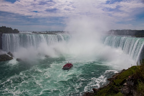 A large, majestic waterfall cascades down a cliff, creating a misty spray that fills the air. In the foreground, a tour boat with a red roof navigates the choppy, turquoise waters below the falls. The scene is framed by lush greenery at the edge of the photo, and the sky above is partly cloudy, adding a dynamic contrast to the scene.