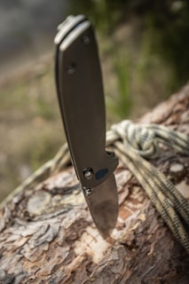 A folding knife is embedded into the surface of a log. The knife handle is metallic with a sleek design. A piece of rope is casually draped over the log, suggesting an outdoor or survival setting. The background appears to be blurred greenery, indicating a natural environment.