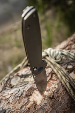 A folding knife is embedded into the surface of a log. The knife handle is metallic with a sleek design. A piece of rope is casually draped over the log, suggesting an outdoor or survival setting. The background appears to be blurred greenery, indicating a natural environment.