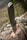 A folding knife is embedded into the surface of a log. The knife handle is metallic with a sleek design. A piece of rope is casually draped over the log, suggesting an outdoor or survival setting. The background appears to be blurred greenery, indicating a natural environment.