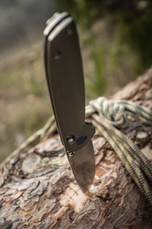 A folding knife is embedded into the surface of a log. The knife handle is metallic with a sleek design. A piece of rope is casually draped over the log, suggesting an outdoor or survival setting. The background appears to be blurred greenery, indicating a natural environment.