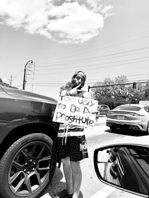 A person stands at the roadside holding a sign with text while wearing a sleeveless shirt and shorts. They are near a parked car, and there are other vehicles on the road. Overhead, the sky is partly cloudy, and power lines can be seen in the background.