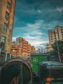 A bustling urban street scene featuring high-rise buildings under a vast blue sky with scattered clouds. The foreground includes a man riding a rickshaw and several vehicles, including a green delivery truck and another rickshaw. Wires and utility poles are visible, adding to the busy urban environment.