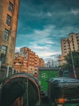 A bustling urban street scene featuring high-rise buildings under a vast blue sky with scattered clouds. The foreground includes a man riding a rickshaw and several vehicles, including a green delivery truck and another rickshaw. Wires and utility poles are visible, adding to the busy urban environment.