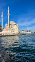 A beautiful mosque with two minarets stands by the waterfront under a clear blue sky. A bridge is visible in the background along with a bustling promenade lined with people and white tents.