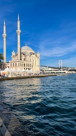 A beautiful mosque with two minarets stands by the waterfront under a clear blue sky. A bridge is visible in the background along with a bustling promenade lined with people and white tents.