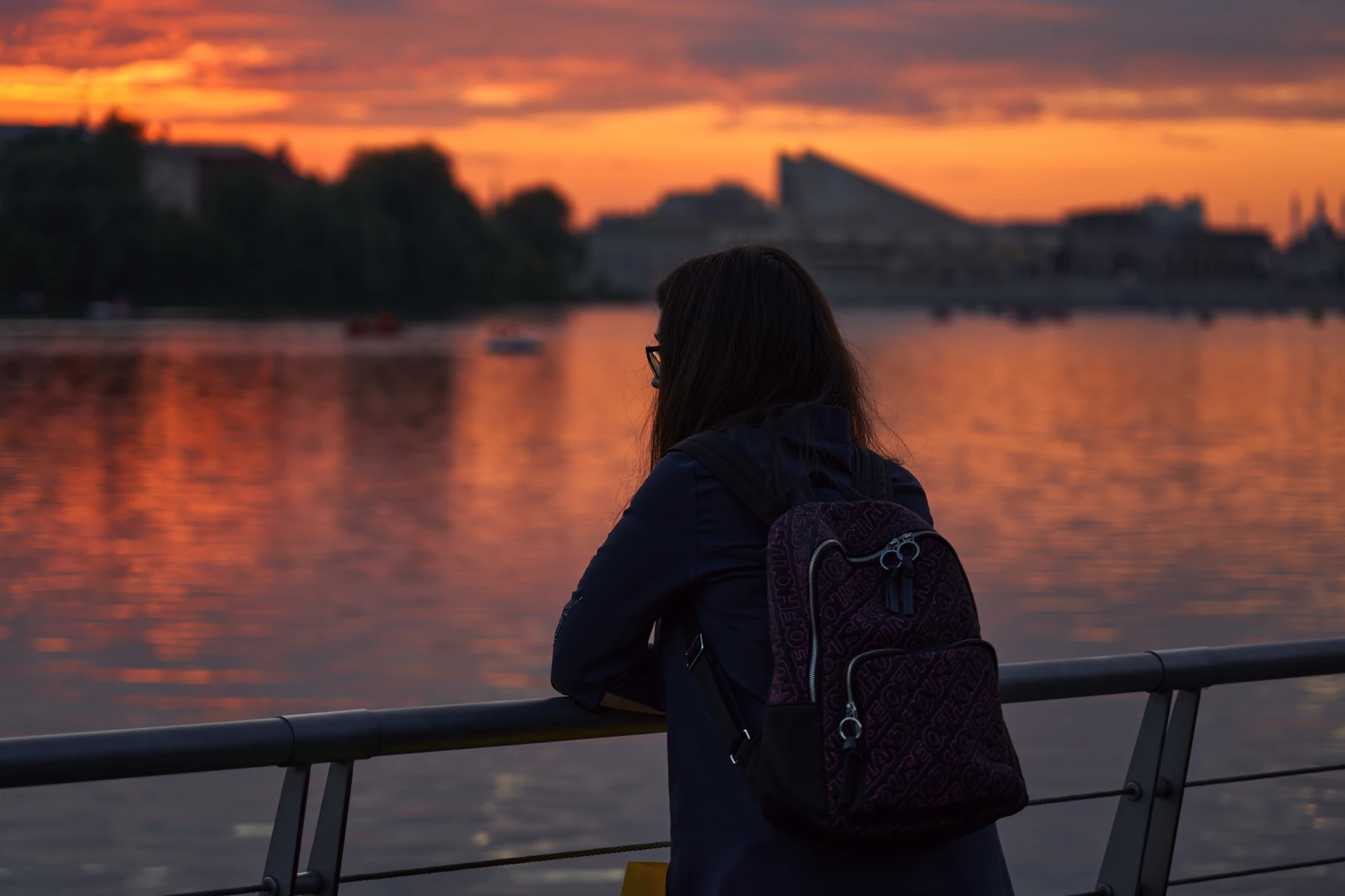 Student with a backpack watching an orange sunset over a river in a city at dusk