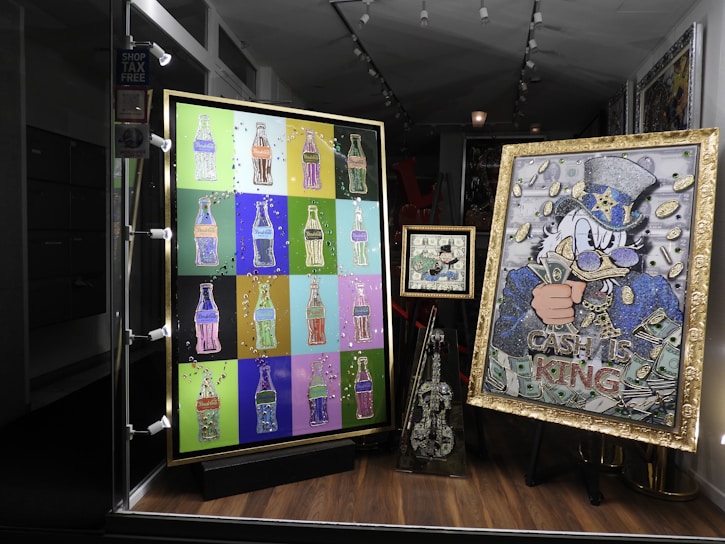 A shop window display featuring colorful pop art. The left artwork consists of multiple bottle-shaped images in various bright colors on a grid. The right artwork features a character in a hat with cash motifs and the words 'Cash is King'. A smaller piece in the center and a decorated violin-like sculpture are also included.