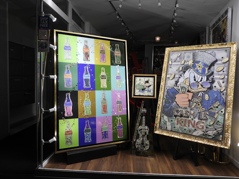 A shop window display featuring colorful pop art. The left artwork consists of multiple bottle-shaped images in various bright colors on a grid. The right artwork features a character in a hat with cash motifs and the words 'Cash is King'. A smaller piece in the center and a decorated violin-like sculpture are also included.