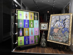 A shop window display featuring colorful pop art. The left artwork consists of multiple bottle-shaped images in various bright colors on a grid. The right artwork features a character in a hat with cash motifs and the words 'Cash is King'. A smaller piece in the center and a decorated violin-like sculpture are also included.