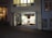 A small art gallery named Donde Art, located on a cobblestone street. The gallery's entrance is illuminated, showcasing various colorful artworks in the window display. The interior is softly lit, and visible artworks include a mix of contemporary styles. The gallery's name and a list of cities are displayed prominently above the window.