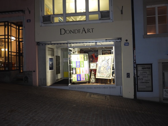 A small art gallery named Donde Art, located on a cobblestone street. The gallery's entrance is illuminated, showcasing various colorful artworks in the window display. The interior is softly lit, and visible artworks include a mix of contemporary styles. The gallery's name and a list of cities are displayed prominently above the window.