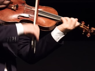 A person is playing the violin, holding the bow in the right hand while the left hand fingers the strings. The violin is classic with a warm wooden tone, and the person is wearing a dark suit with a white shirt.