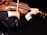 A person is playing the violin, holding the bow in the right hand while the left hand fingers the strings. The violin is classic with a warm wooden tone, and the person is wearing a dark suit with a white shirt.