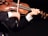 A person is playing the violin, holding the bow in the right hand while the left hand fingers the strings. The violin is classic with a warm wooden tone, and the person is wearing a dark suit with a white shirt.