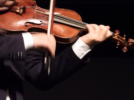 A person is playing the violin, holding the bow in the right hand while the left hand fingers the strings. The violin is classic with a warm wooden tone, and the person is wearing a dark suit with a white shirt.