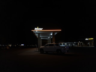 A dimly lit car wash at night features a parked white truck under the illuminated canopy. The sign above reads 'Mister' with a yellow glow, while another sign on a nearby building displays 'Mister Car Wash'. The surrounding area is dark with few visible details.