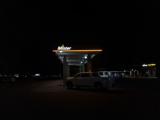 A dimly lit car wash at night features a parked white truck under the illuminated canopy. The sign above reads 'Mister' with a yellow glow, while another sign on a nearby building displays 'Mister Car Wash'. The surrounding area is dark with few visible details.