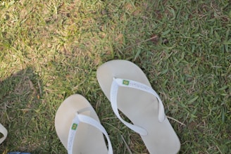 Two white flip-flops are positioned on a patch of grass. The grass is lush and green, with a few brown spots indicating slight dryness. The footwear is labeled with a small flag emblem near the strap junction.