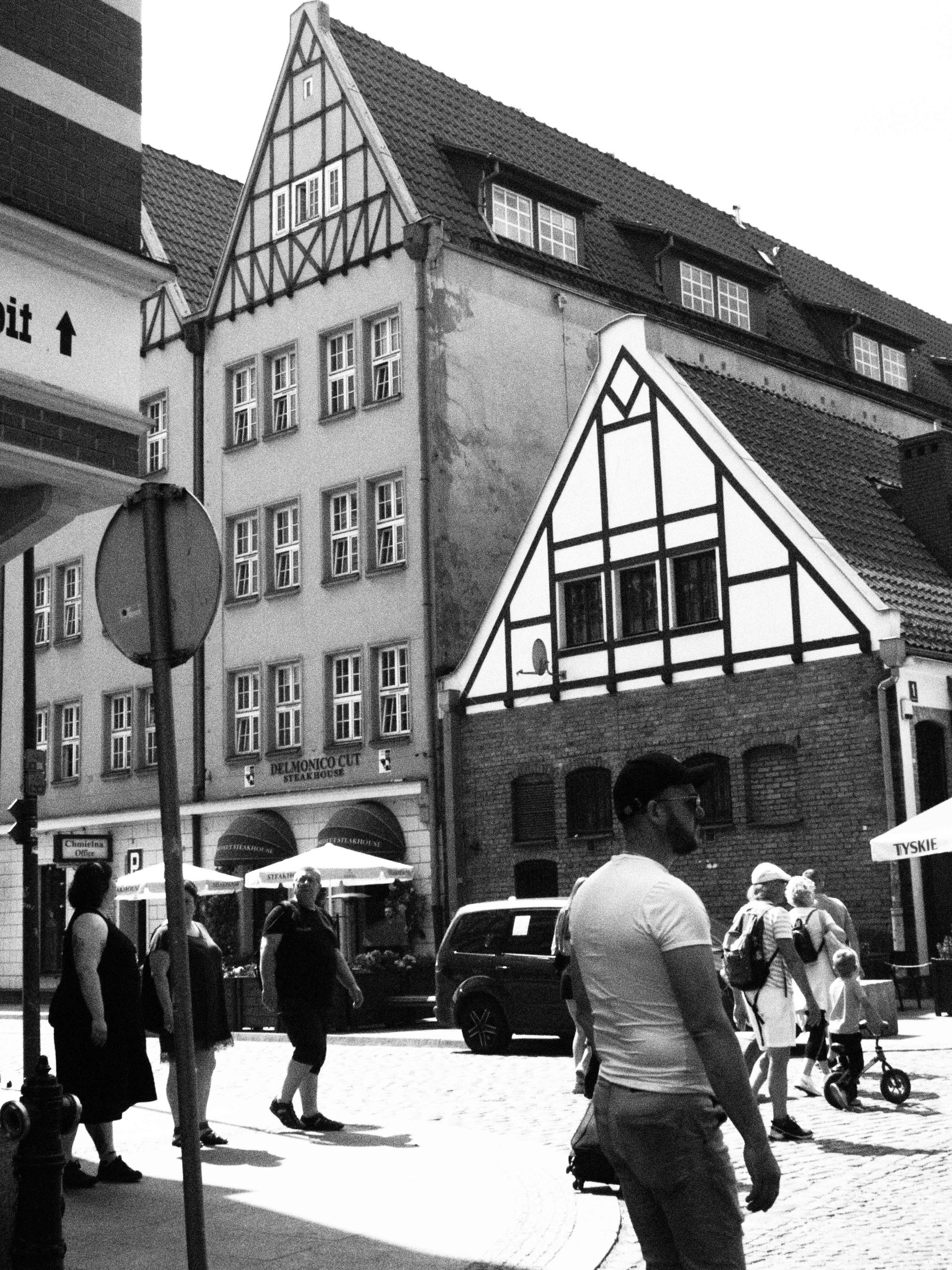 Monochrome street scene in a historic European town with half-timbered buildings, pedestrians, and a parked vehicle.