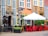 A quaint street scene featuring a rustic brick building with tall windows and decorative architectural elements. A wooden statue stands near the entrance of a venue, and a canopy shades an outdoor seating area with bright red chairs around a black table. Plants and a decorative arch enhance the cozy atmosphere.