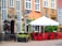 A quaint street scene featuring a rustic brick building with tall windows and decorative architectural elements. A wooden statue stands near the entrance of a venue, and a canopy shades an outdoor seating area with bright red chairs around a black table. Plants and a decorative arch enhance the cozy atmosphere.