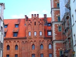 A historic brick building with Gothic architectural features, including pointed arches and a red terracotta roof. The facade includes multiple windows with decorative frames, while scaffolding is visible on one side of the structure under renovation.