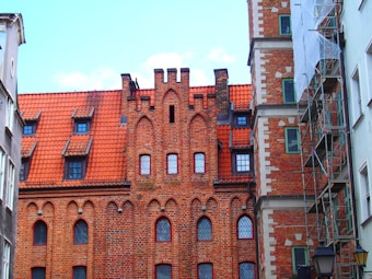 A historic brick building with Gothic architectural features, including pointed arches and a red terracotta roof. The facade includes multiple windows with decorative frames, while scaffolding is visible on one side of the structure under renovation.