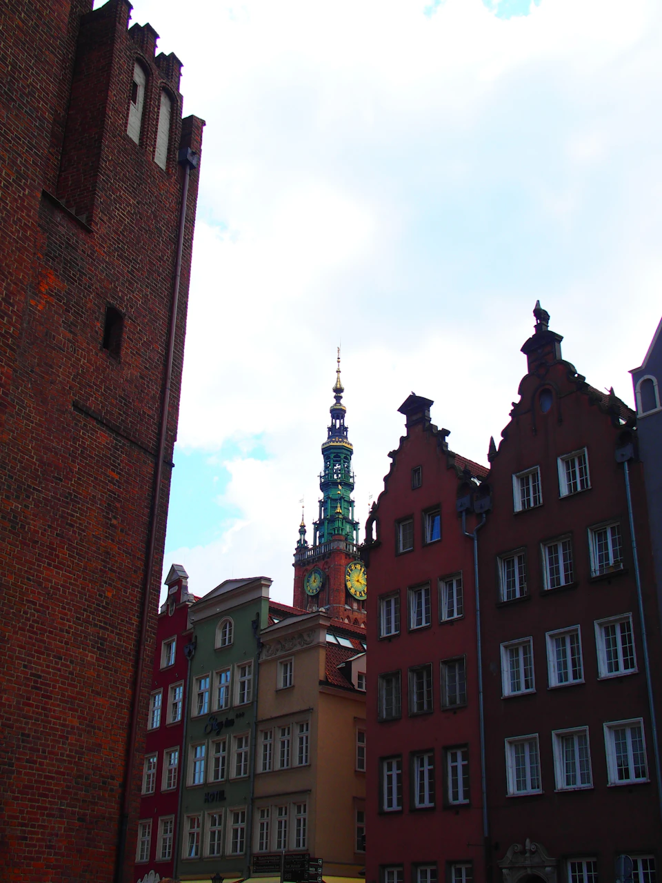 The image depicts a cityscape with a tall brick tower featuring a clock face, standing out against the sky. The tower is part of a larger building that also has a clock on its facade. The surrounding buildings are painted in various colors and have unique architectural styles, creating a vibrant and diverse urban scene.