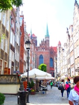A cobblestone street lined with historic buildings leads toward a large brick cathedral with tall spires. People are walking along the street, some browsing through outdoor market stalls covered with white umbrellas. Flower pots and greenery add color to the scene, and there is a streetlamp on the side.